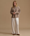 A woman stands against a warm beige studio background wearing a taupe quarter-zip knit sweater and wide-leg cream pants with brown shoes.
