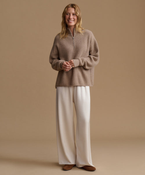 A woman stands against a warm beige studio background wearing a taupe quarter-zip knit sweater and wide-leg cream pants with brown shoes.