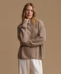 A woman wearing a beige ribbed knit half-zip sweater and cream trousers, posing against a solid beige studio background.