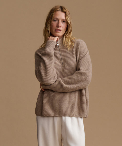 A woman wearing a beige ribbed knit half-zip sweater and cream trousers, posing against a solid beige studio background.
