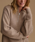 A woman wearing a chunky ribbed knit half-zip sweater in a warm taupe-beige color, shown from the chest up against a solid beige background.