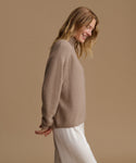 A woman wearing a beige ribbed knit sweater and white trousers, shown from a side profile against a solid beige studio background.