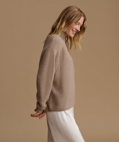 A woman wearing a beige ribbed knit sweater and white trousers, shown from a side profile against a solid beige studio background.