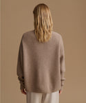 A woman viewed from behind wearing a taupe ribbed knit sweater and light beige trousers, set against a solid warm beige studio background.