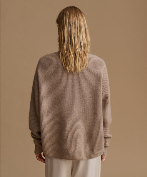 A woman viewed from behind wearing a taupe ribbed knit sweater and light beige trousers, set against a solid warm beige studio background.