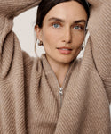 A woman with light eyes and brown hair wears the Cashmere Half Zip in undyed stone. She poses with arms raised, looking at the camera, wearing minimal makeup and silver hoop earrings.