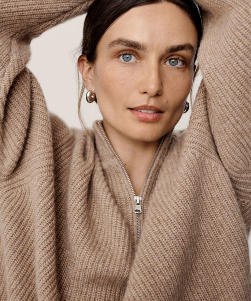 A woman with light eyes and brown hair wears the Cashmere Half Zip in undyed stone. She poses with arms raised, looking at the camera, wearing minimal makeup and silver hoop earrings.