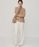 A woman stands against a plain background, smiling with one hand partly covering her mouth. She wears the undyed stone Cashmere Half Zip sweater, white wide-leg pants, open-toed shoes, and carries a beige shoulder bag.