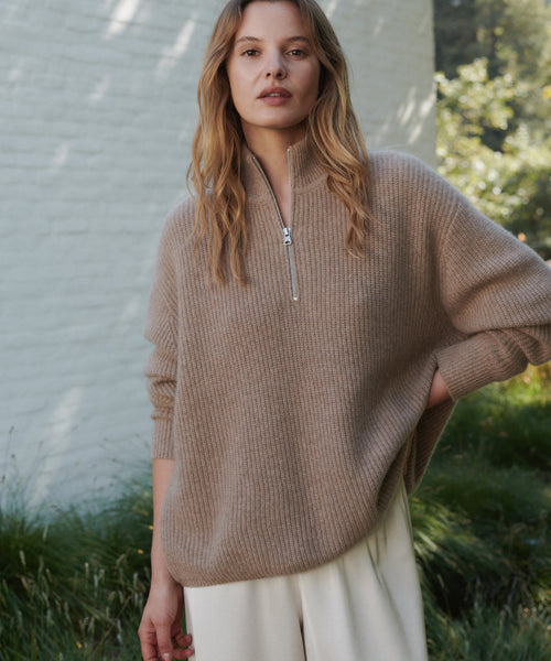 A woman wearing a beige ribbed knit half-zip sweater wpaired with cream wide-leg trousers, standing outdoors in front of a white brick wall and green foliage.