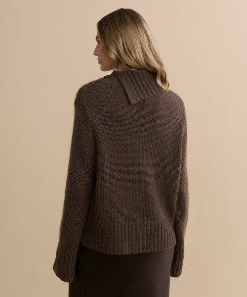 A woman with light brown hair is seen from the back, wearing the Cashmere Jackie Sweater—an elegant brown knit with a high, uneven collar and ribbed edges—paired with a dark skirt against a beige backdrop.