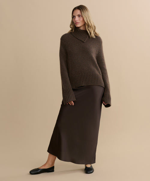 A woman stands against a beige background wearing the Cashmere Jackie Sweater in brown, featuring button-shoulder details, paired with a long dark brown skirt and black flats. She looks to the side with one hand by her side and the other slightly bent.