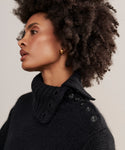 A woman with short, natural curly hair wears the Cashmere Jackie Sweater—a chunky black luxury knit with a buttoned high collar. She has gold hoop earrings and looks to the left with a neutral expression.