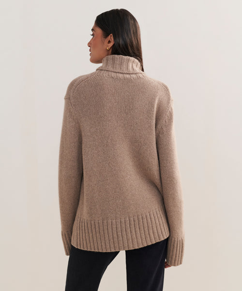 A person with long dark hair faces away from the camera, wearing the oversized Cashmere Jet Turtleneck sweater and dark pants, set against a plain light background.