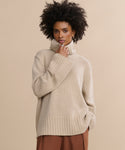 A woman with curly hair, wearing the Cashmere Jet Turtleneck and brown pants, stands against a plain beige background with a thoughtful expression.