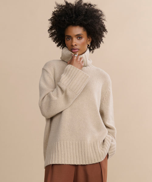 A woman with curly hair, wearing the Cashmere Jet Turtleneck and brown pants, stands against a plain beige background with a thoughtful expression.