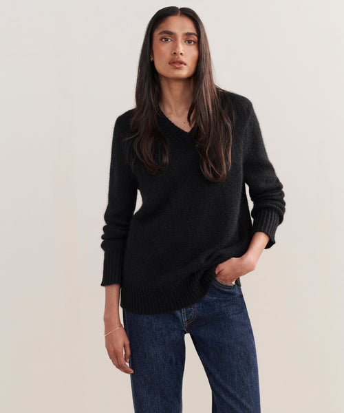 A woman with long dark hair wears the Cashmere Kate V-Neck sweater in black and blue jeans, standing against a light-colored background with a neutral expression.