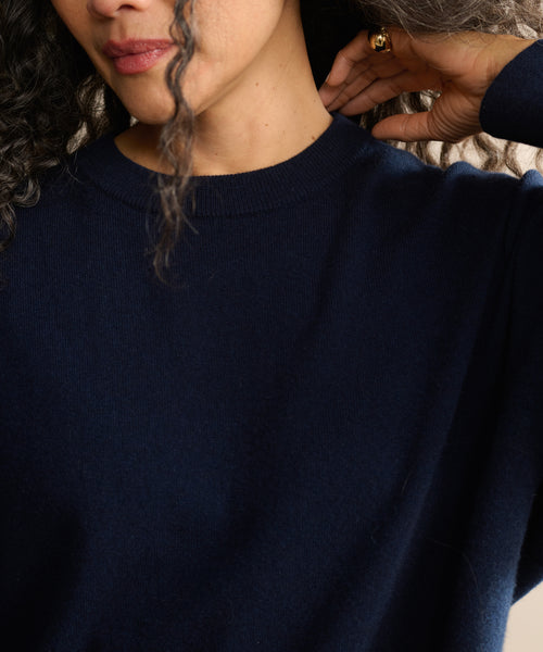 A person with curly hair wears the Cashmere Marina Crewneck in dark navy blue, gently touching their neck. Only the lower face and upper torso are visible.