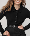 A woman with wavy brown hair smiles, wearing the Murphy Cardigan and black belted trousers, with a gold ring. Her hand rests on her waist against a plain light background.