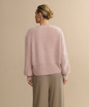 A person with blonde hair in a low bun is shown from the back, wearing a Cashmere Peyton Cardigan in soft pink with long sleeves and loose beige trousers, standing against a plain beige background.