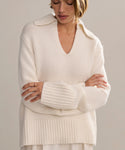 A woman stands with arms crossed, looking downward in the Cashmere Phoebe Polo—an oversized ivory sweater with a deep V-neck and wide collar, crafted from soft jersey-knit fabric that complements the neutral backdrop.