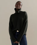 A person models the Cashmere Porter Turtleneck with blue jeans, standing against a beige background and looking confidently at the camera with hands clasped.