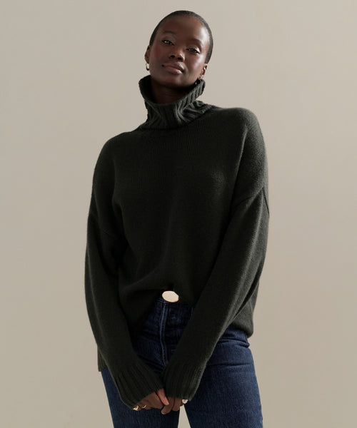 A person models the Cashmere Porter Turtleneck with blue jeans, standing against a beige background and looking confidently at the camera with hands clasped.