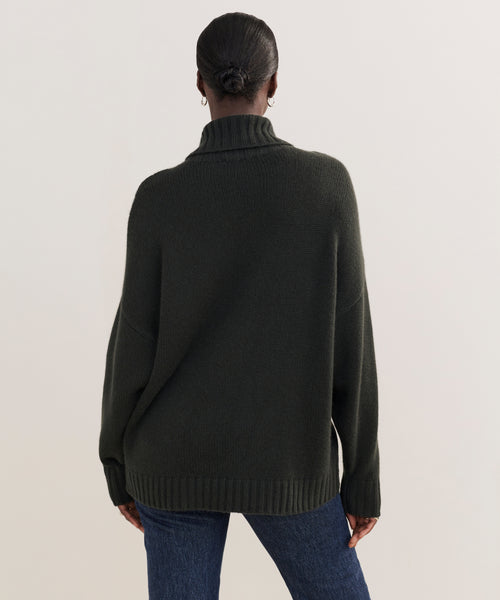 A person stands facing away from the camera, wearing the Cashmere Porter Turtleneck in dark green and blue jeans. Their hair is styled in a neat bun, set against a plain, light-colored background.
