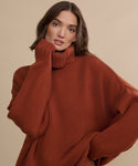 A woman with wavy brown hair wears the Cashmere Porter Turtleneck in rust, standing against a beige background. One hand touches the collar as she poses with a relaxed expression.