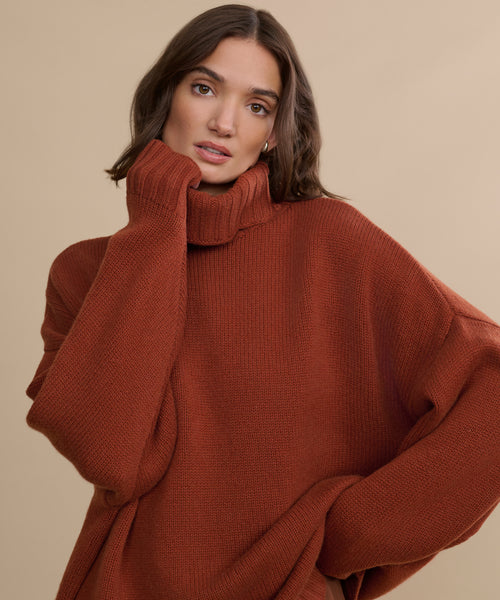 A woman with wavy brown hair wears the Cashmere Porter Turtleneck in rust, standing against a beige background. One hand touches the collar as she poses with a relaxed expression.