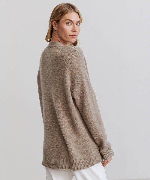 A woman with blonde hair, dressed in a long beige Cashmere Spencer Cardigan and white pants, stands against a plain light background, looking over her shoulder toward the camera.
