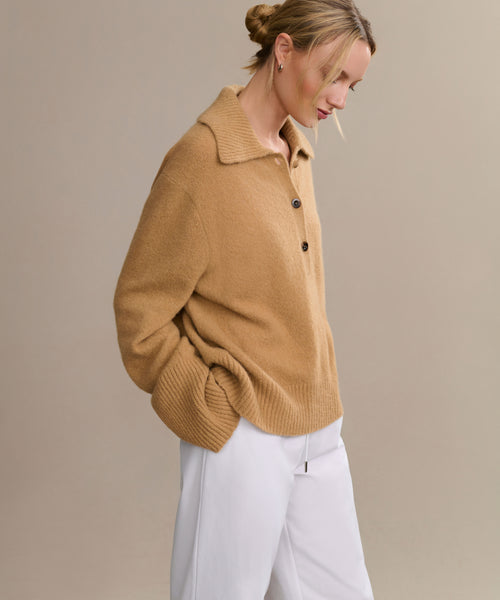 A woman with blonde hair in a low bun wears the Cashmere Stevie Polo with an oversized collar and white pants, standing against a beige background, looking down with one hand in her pocket.