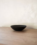 A minimal scene features the Cast Iron Bowl by Blackcreek Mercantile, crafted in the USA. The simple round bowl sits on a light wood surface against an off-white wall, highlighted by soft lighting.
