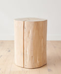 The Cedar Stump, made from reclaimed cedarwood, features visible grain and a vertical crack. This handcrafted piece serves as a rustic stool or side table, adding unique natural charm to any space.