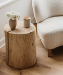 A cozy scene with a natural accent: the Cedar Stump, handcrafted from reclaimed cedarwood, serves as a side table holding a small vase of white flowers and a lit candle next to a white textured armchair on a wooden floor.