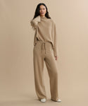 A woman with long dark hair stands against a beige background wearing the Chalet Pant—a loose, light brown felted wool loungewear pant with a tie waist—and a matching sweater. She pairs them with white shoes and touches her neck.