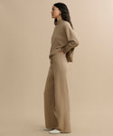 A woman in profile stands against a beige background, wearing the loose-fitting Chalet Pant loungewear set with wide legs, her hands in her pockets and white shoes peeking out.
