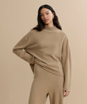 A woman with long dark hair models the Chalet Sweater, a beige, loose-fitting loungewear set, standing against a neutral background with one hand on her hip and a subtle smile.