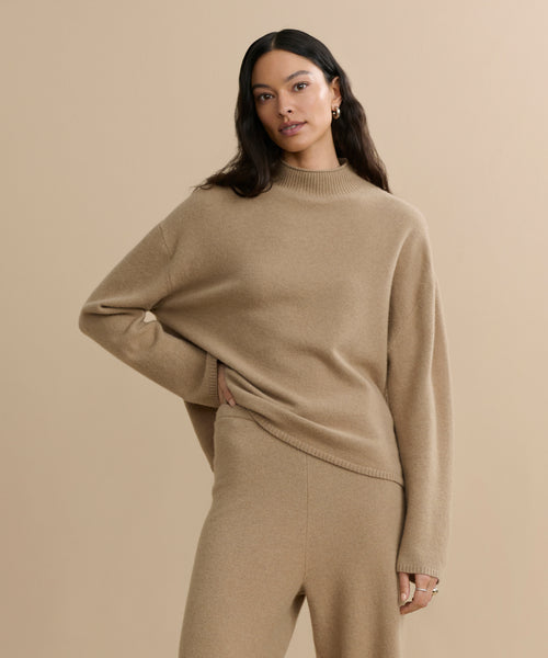 A woman with long dark hair models the Chalet Sweater, a beige, loose-fitting loungewear set, standing against a neutral background with one hand on her hip and a subtle smile.