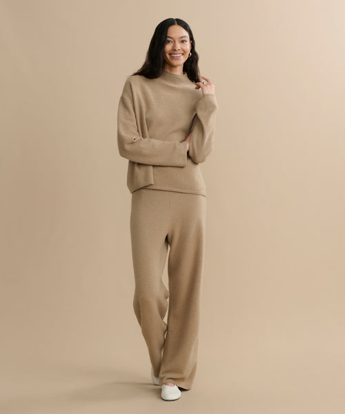 A woman stands against a beige background, smiling in the Chalet Sweater loungewear set—a felted wool blend with wide-leg pants—paired with white slip-on shoes.