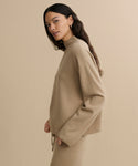 A woman with long dark hair models the loose-fitting Chalet Sweater in beige with matching pants, standing against a light beige background and looking toward the camera.