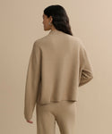 A person with long, wavy dark hair wears a loose Chalet Sweater and matching pants from a felted wool blend loungewear set, facing away from the camera against a plain beige background.