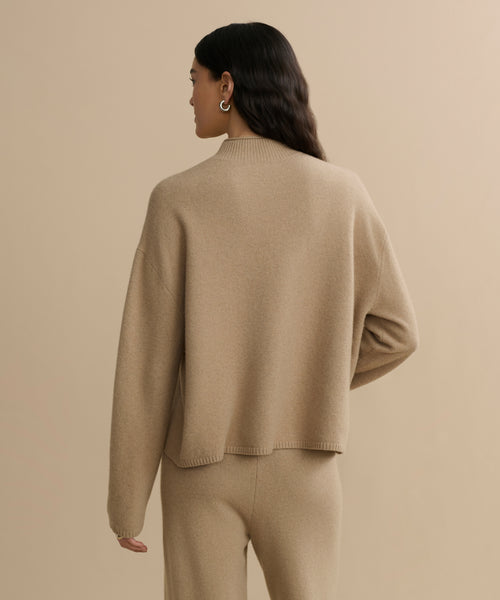 A person with long, wavy dark hair wears a loose Chalet Sweater and matching pants from a felted wool blend loungewear set, facing away from the camera against a plain beige background.