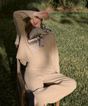 A woman in the Chalet Sweater loungewear set sits on a wooden chair outdoors, smiling and shielding her eyes from the sun. She wears a patterned scarf over her shoulders and is surrounded by green grass.