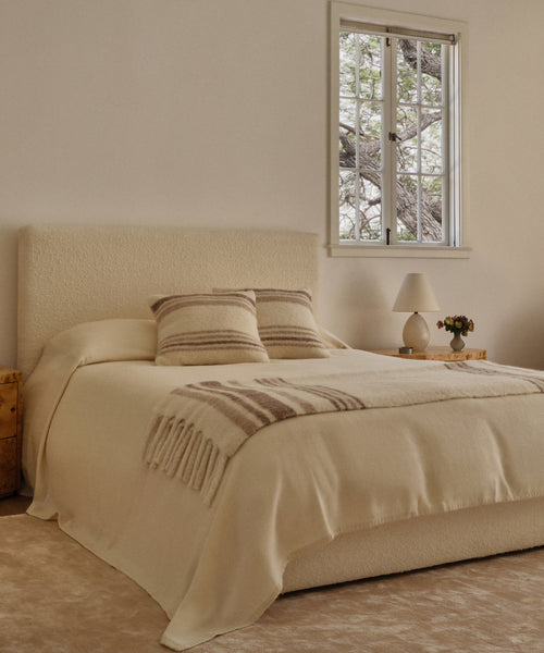 A cozy bedroom with a cream bed featuring the Chateau Alpaca Coverlet, two striped pillows, a folded throw, wooden nightstands, and a sunlit window. One nightstand displays a lamp, vase, and small plant.