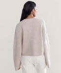 A woman with long dark hair, seen from behind, wears a loose Chelsea Cardigan in oatmeal with high-waisted white jeans against a simple light background.
