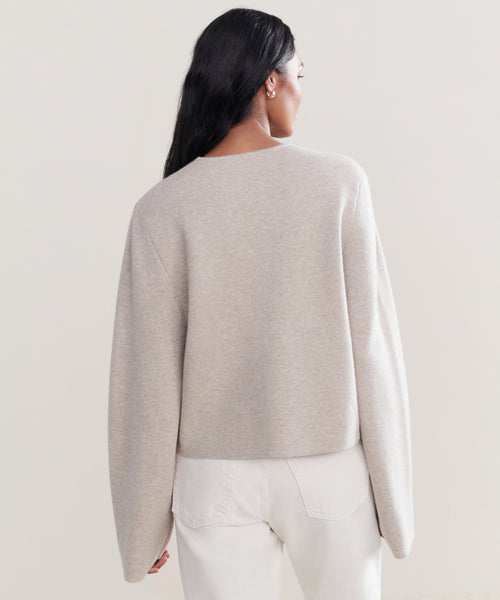 A woman with long dark hair, seen from behind, wears a loose Chelsea Cardigan in oatmeal with high-waisted white jeans against a simple light background.