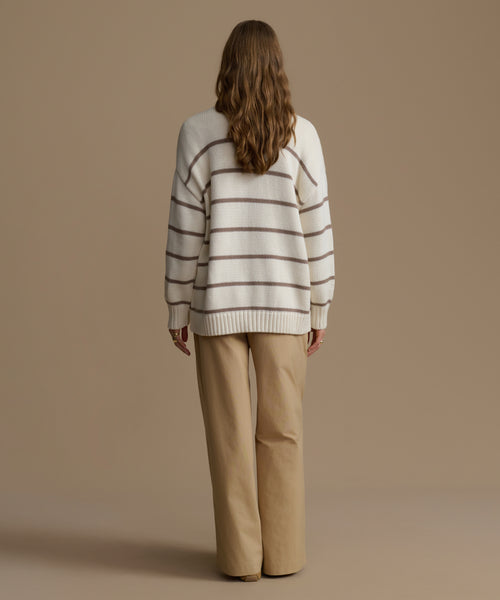 Person wearing a striped sweater and beige pants against a brown background