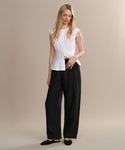 A woman with long blonde hair wears a sleeveless white blouse, loose black pants, and the Clara Mule shoes. She stands against a beige background, looking slightly down with one hand in her pocket.