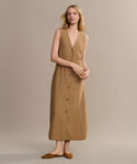 A woman with light hair stands against a beige background, wearing a sleeveless tan dress with front buttons and the Clara Mule shoes in cognac. She appears relaxed, showcasing the Italian-made Clara Mule.