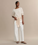 A woman stands against a beige background wearing the Claude Long-Sleeve Tee in cream and tan stripes, paired with white wide-leg pants, sandals, and a matching striped sweater tied at her waist.
